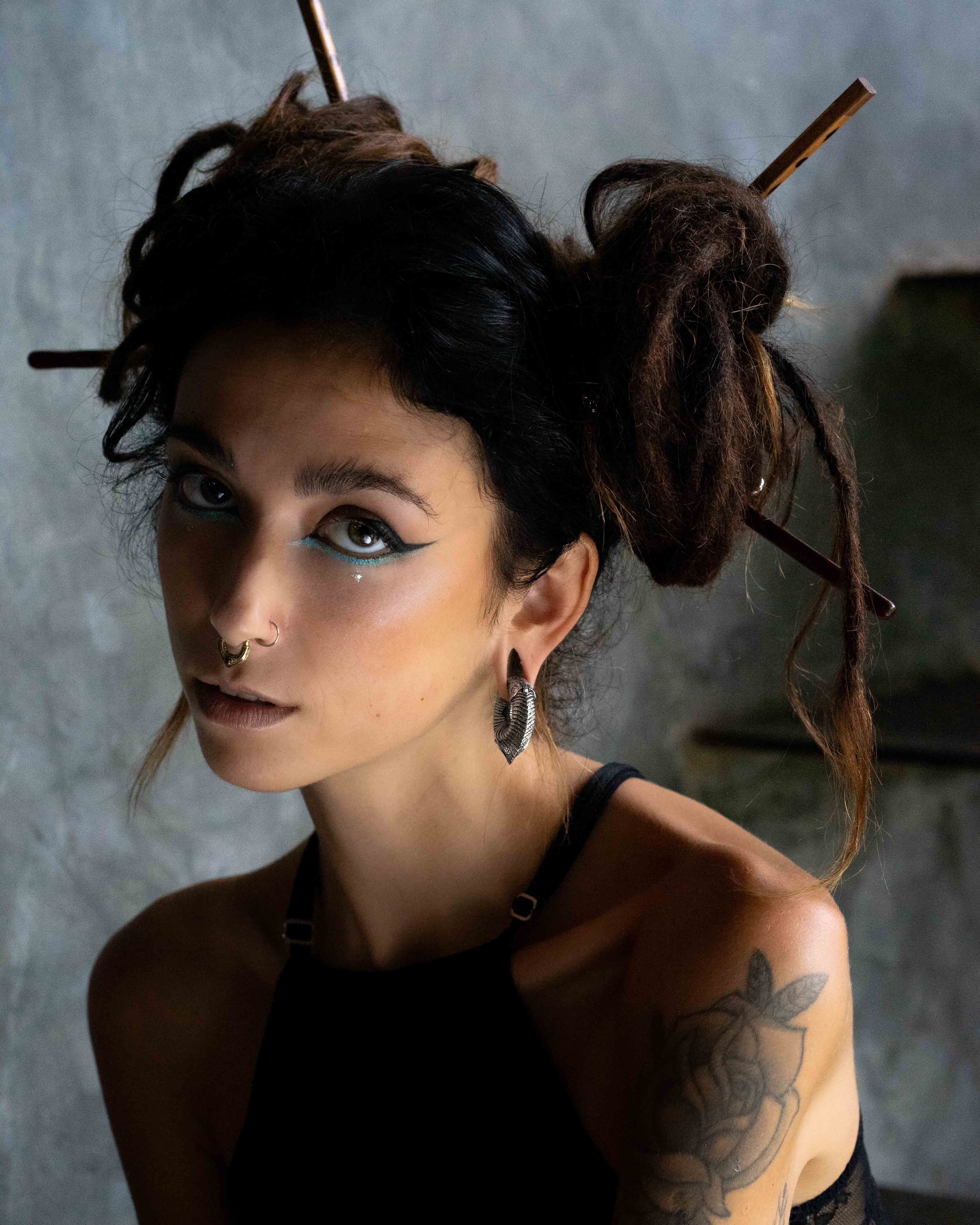 A woman with dark hair styled in buns, adorned with two sticks, gazes at the camera. She wears winged eyeliner, a nose ring, and heavy silver hoop earrings identified as the MARVDASHT Medium Cyborg Clicker Ear Weights in Silver Brass by Kaya Movement. She has on a black tank top and displays a flower tattoo on her upper arm. The background is a textured gray wall.