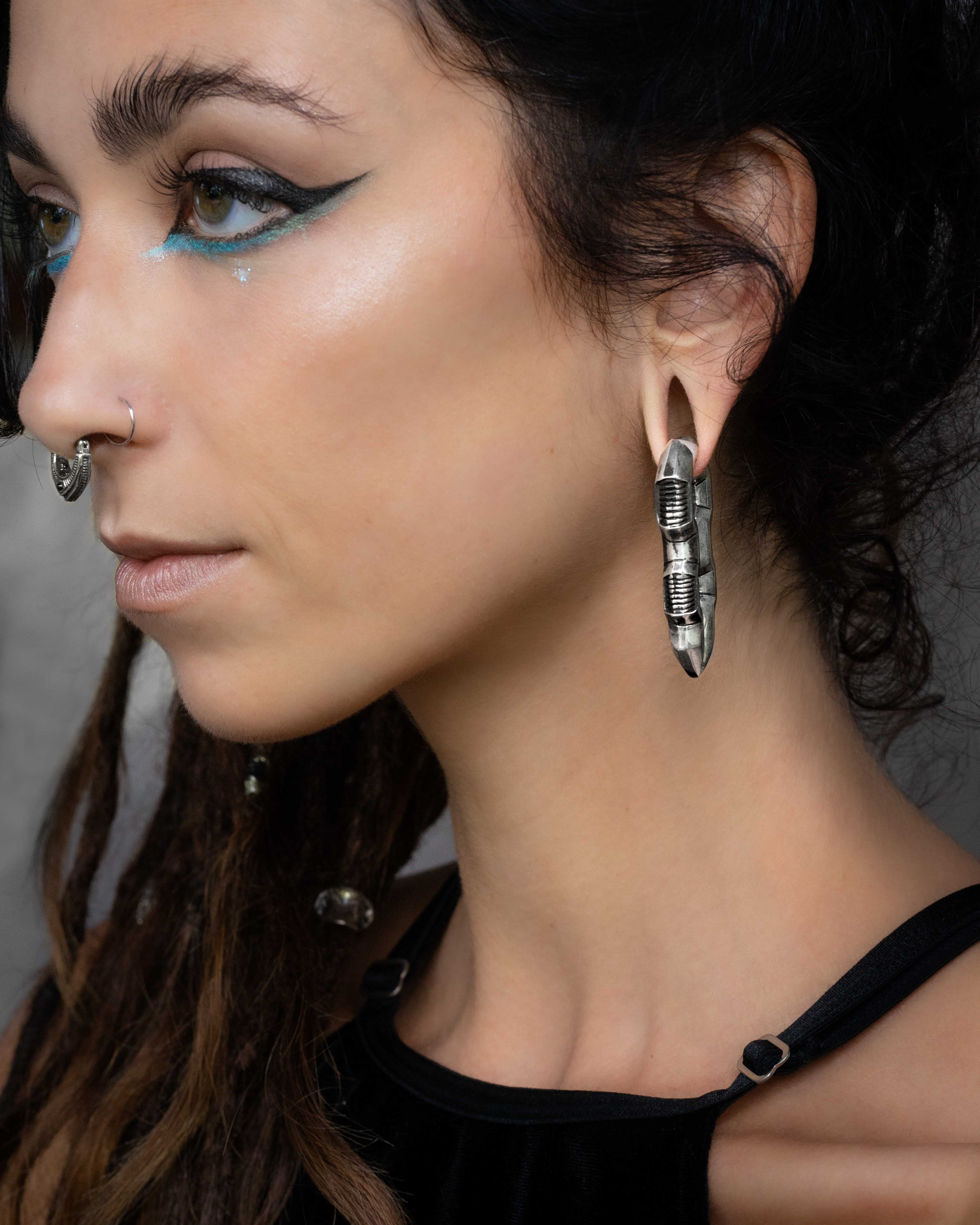 A person with long dark hair and an intricate nose ring is wearing bold silver hoop earrings and dramatic aqua blue eyeliner. They are dressed in a black top with metal ring accents and post-apocalyptic body jewelry, including the unique Kaya Movement TECHRE Futuristic Hexagon Keyhole Ear Weights in Silver (0 gauge). The neutral background highlights their accessories and makeup.