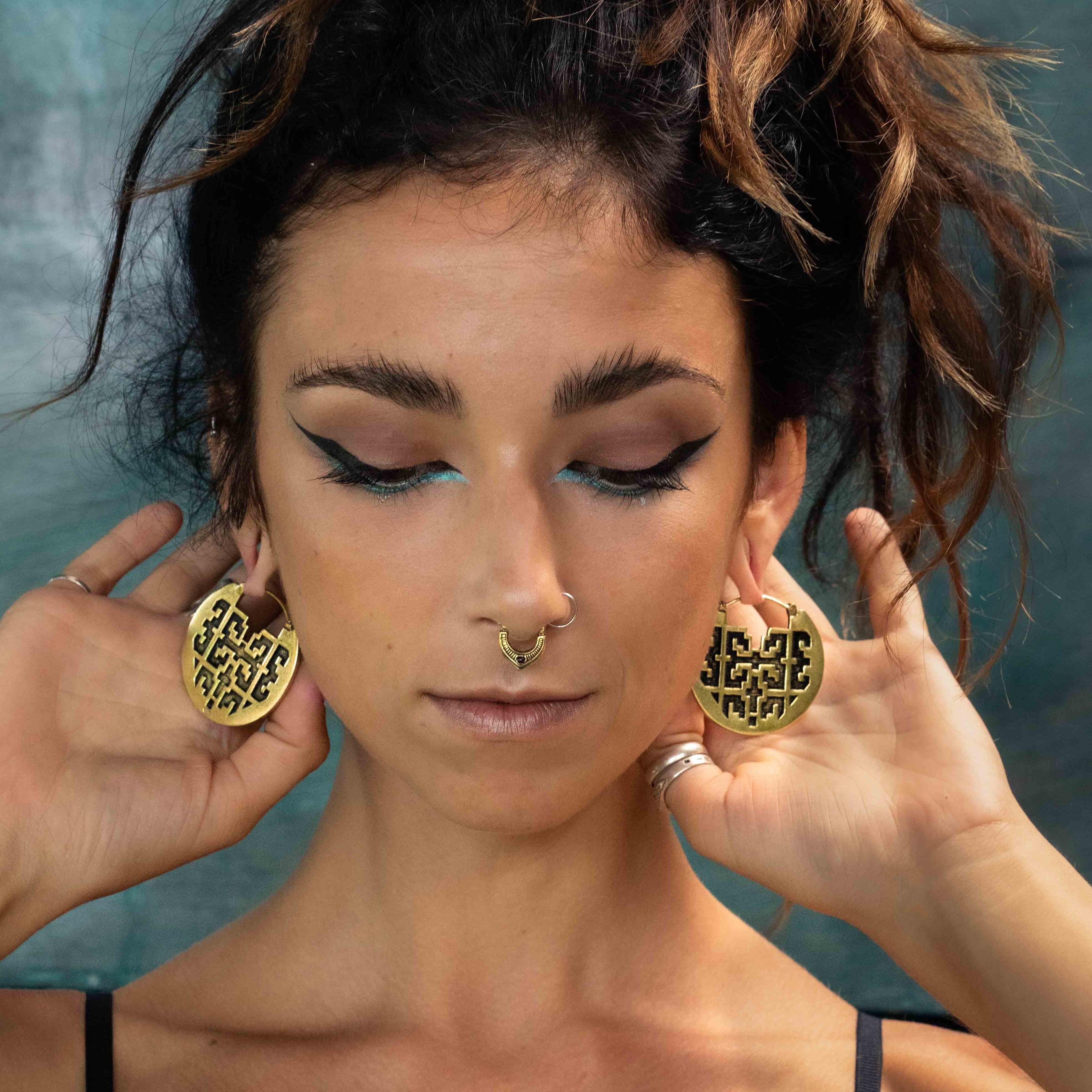 A person with dark makeup and winged eyeliner holds up the large CONIBOS Tribal Shipibo Hoop Disc Earrings in Gold from Kaya Movement against their ears. They have a nose ring, and their hair is loosely tied up, with some loose strands framing their face. The boho body jewelry stands out against the muted, textured background.
