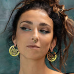 A woman with dark, curly hair tied up is looking to the side, showcasing bold makeup with winged eyeliner and pale blue underlining. She wears large CONIBOS Tribal Shipibo Hoop Disc Earrings in Gold by Kaya Movement and a septum ring, set against a blurred teal gradient background.
