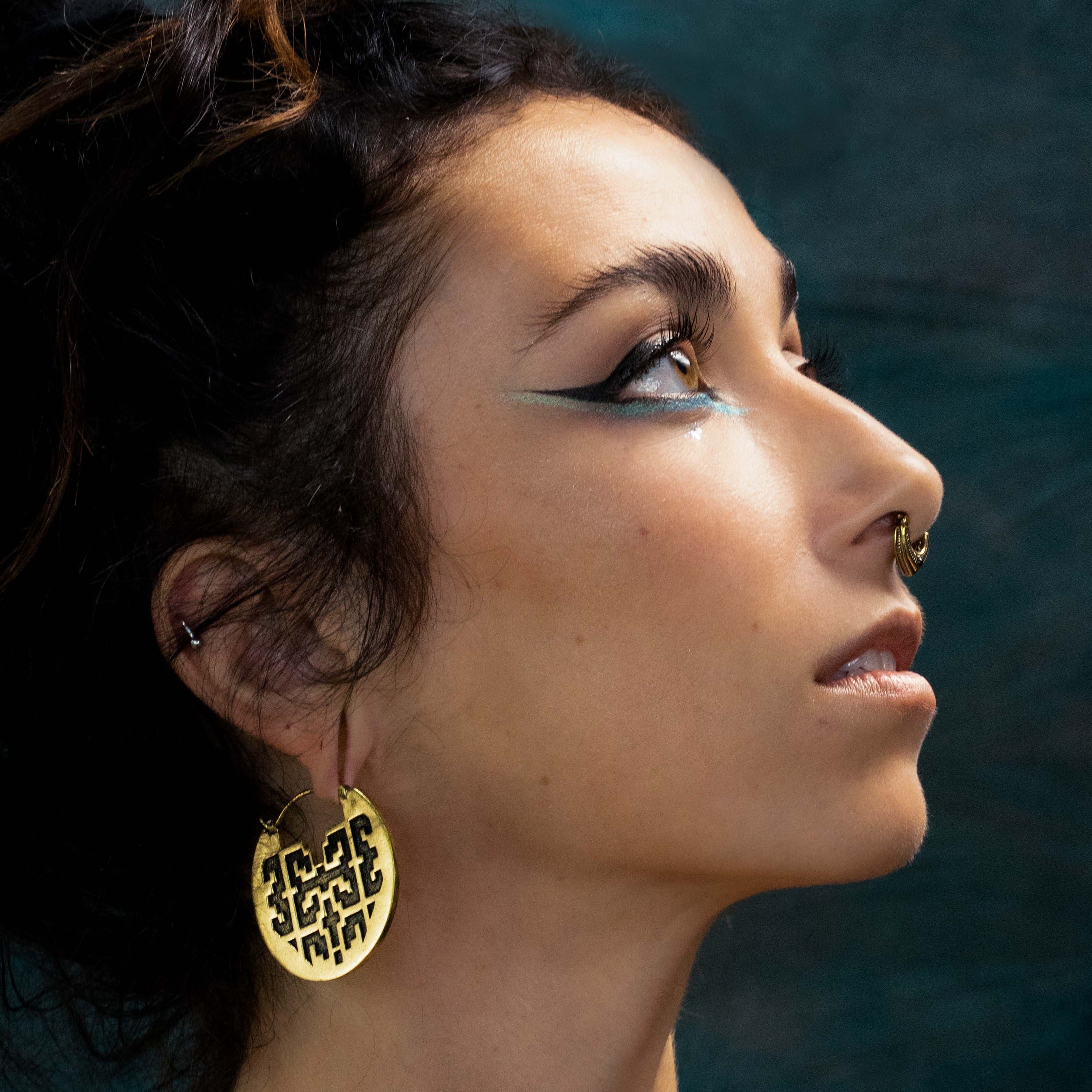 Close-up profile of a person with dark hair styled in a bun. They have dramatic teal and black eye makeup, a nose ring, and are wearing Kaya Movement's CONIBOS Tribal Shipibo Hoop Disc Earrings in Gold (16 gauge), which feature intricate designs. The background is blurred and dark, emphasizing their facial features and accessories.