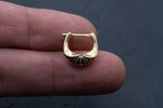 A person's fingers holding the DIARA Biomechanical Clicker Hoop Septum Nose Ring by Kaya Movement against a dark background. The unisex, 16-gauge gold nose ring features a ridged design pattern at the bottom.