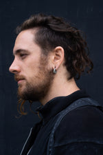 A person with long, wavy hair tied back and a beard is shown in profile view against a dark background. They are wearing a dark-colored top and have Kaya Movement's AARO Cyborg Clicker Hoop Ear Weights in 925 Silver, 6 gauge, in their stretched ears and a matching septum piercing.