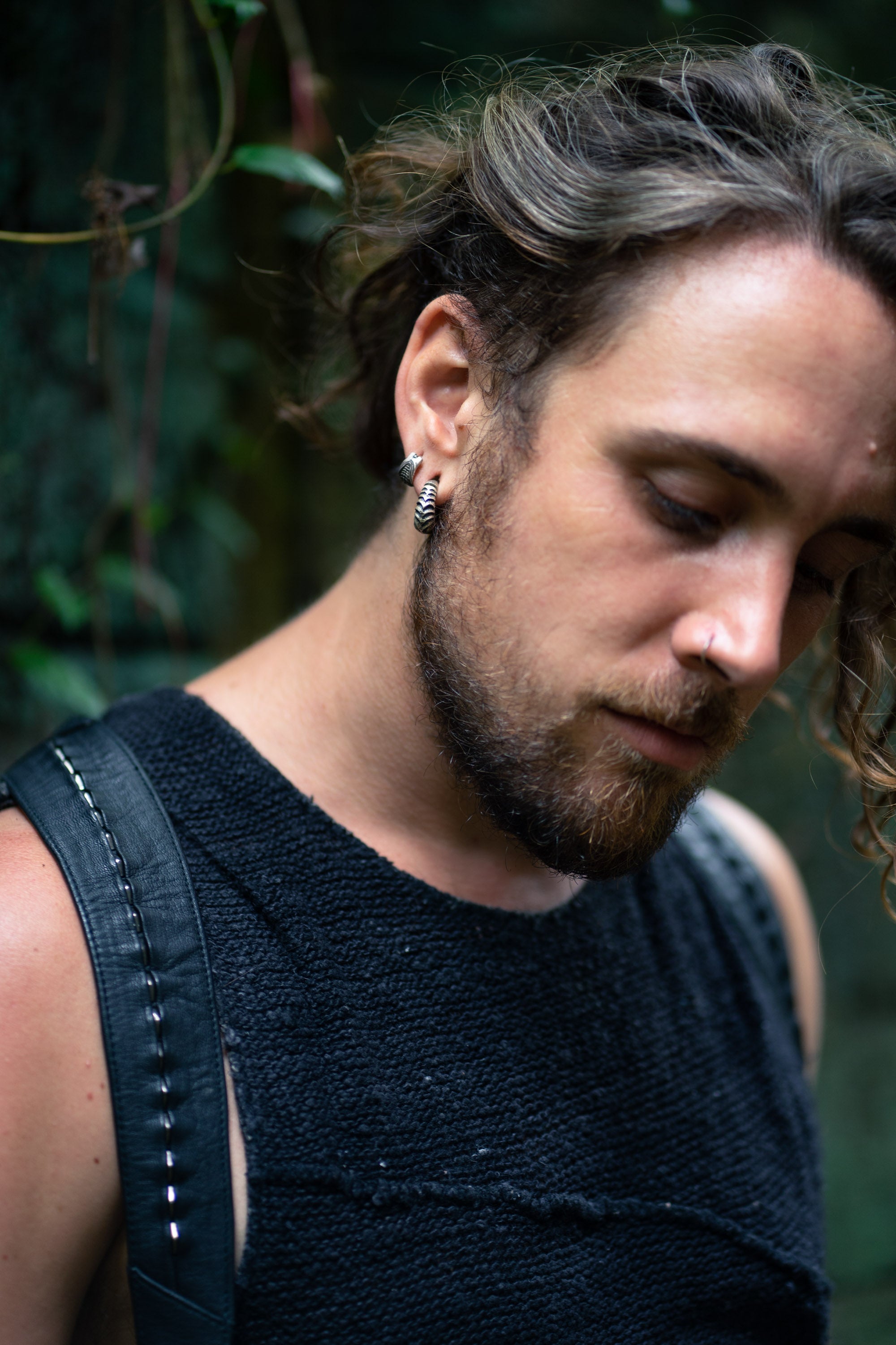 A person with wavy, long hair and facial hair looks down pensively. The individual is wearing a black textured sleeveless top and has an earring, a nose ring, and stretched ears adorned with AARO Cyborg Clicker Hoop Ear or Septum Weights in 925 Silver from Kaya Movement. The backdrop is blurred greenery, suggesting an outdoor setting.