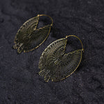 Introducing the CIRCUIT Oversized Visionary Disc Hoop Earrings in Gold by Kaya Movement. These 16-gauge earrings boast an intricately designed maze-like pattern in a circular form. Each segment is delicately cut out to create an eye-catching geometric design, perfect for making a boho statement. Pictured on a textured, dark surface, these gold-colored earrings are a true standout accessory.