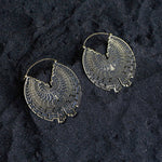 A pair of Kaya Movement's CIRCUIT Oversized Visionary Disc Hoop Earrings in Gold, crafted with an intricate 16-gauge radial, labyrinthine pattern, lie on a textured dark sandy surface. These boho statement earrings are shaped with precision and feature a maze-like geometric design.