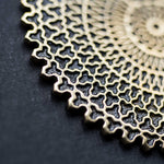 Close-up of the Kaya Movement's VORTEX Tribal Oversized Mandala Disc Hoop Earrings in Gold against a dark gray background. The 16 gauge earrings feature interlocking shapes, forming a detailed and symmetrical mesh with a lace-like appearance. The lighting accentuates the shine and intricate texture of the metallic design.