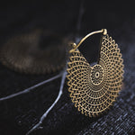 A close-up of the VORTEX Tribal Oversized Mandala Disc Hoop Earrings in Gold by Kaya Movement reveals an intricately designed, geometric, lattice-like pattern resting on a dark surface. Another similar earring is faintly visible in the blurred background, emphasizing the craftsmanship and detail of these boho statement earrings.