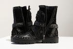 A pair of rugged design black, mid-calf leather boots featuring studded accents, chunky soles, and half-zipped fronts. Both boots have a textured finish and decorative straps with metal embellishments. These unisex MAGMA Wasteland Studded Leather Ankle Combat Boots by Kaya Movement are displayed on a white surface.