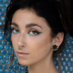 Close-up of a person with dark hair against a textured blue background. They have bold eye makeup featuring black eyeliner and a touch of turquoise, stretched ears adorned with HEXA Futuristic Geometry Hexagon Flesh Tunnels in Gold by Kaya Movement, and a nose ring. Their expression is neutral and confident.
