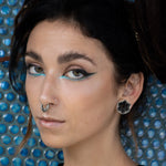 A person with dark hair styled in an updo is adorned with bold blue and black eye makeup, a nose ring, an ear cuff, and body jewelry. Among the accessories is the Kaya Movement HEXA Futuristic Geometry Hexagon Flesh Tunnels in Silver (12mm size) visible in their earlobe. The background features a blue textured pattern, complementing the individual's calm and serious expression.