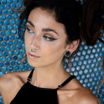 A person with dark hair styled in an updo, donning a black top with spaghetti straps, showcases a facial piercing and Kaya Movement's HEXA Futuristic Geometry Hexagon Flesh Tunnels in Silver. They accentuate their features with bold eyeliner and turquoise eye makeup. The background boasts a textured blue honeycomb-like pattern.