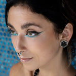A close-up of a person with dark hair, wearing striking turquoise and black winged eyeliner and a nose ring, displays the HEXA Futuristic Geometry Hexagon Flesh Tunnels in Silver by Kaya Movement. The background features a blue pattern. They have a calm expression and their head is slightly tilted to the right.
