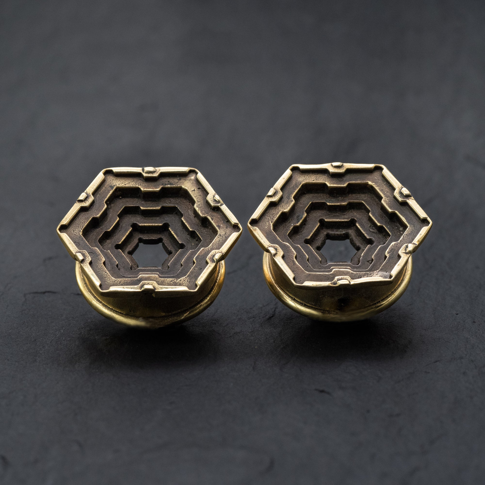 A pair of Kaya Movement's HEXA Futuristic Geometry Hexagon Flesh Tunnels in gold, available in sizes ranging from 12mm to 24mm, featuring an intricate and layered geometric design. Reminiscent of visionary AI plug earrings, the flesh tunnels are positioned side by side on a dark, textured background.