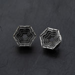 A pair of HEXA Futuristic Geometry Hexagon Flesh Tunnels in Silver from Kaya Movement rests on a black, textured surface. These body jewelry pieces feature intricate, layered designs that create a geometric, 3D appearance. Each layer descends inward toward the center, forming a visually striking pattern. Available in sizes ranging from 12mm to 24mm.