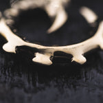 A close-up shot features the GIGER Biomechanical Open Ended Hoop Ear Weights in Gold from Kaya Movement, resembling an ornate, metallic object with spiked ring-like or circular gear qualities. Resting on a dark, textured surface, the ear weights have a polished, reflective finish and are slightly out of focus to highlight their curved and pointed features.