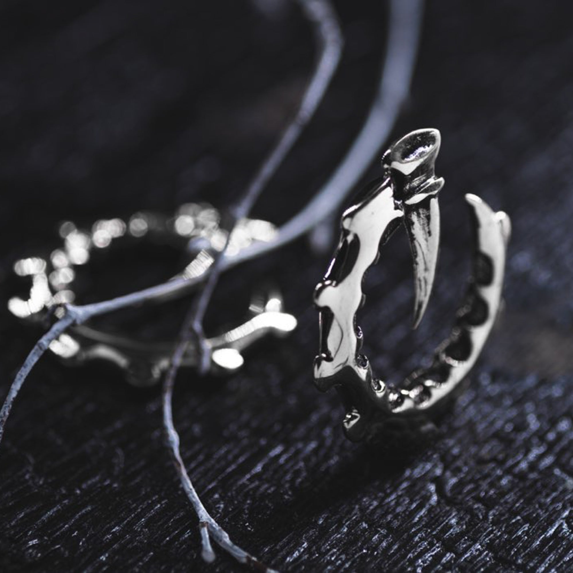 The GIGER Biomechanical Open Ended Hoop Ear Weights in Silver 925 by Kaya Movement are showcased on a dark, textured surface. Thin, string-like elements partially surround the ear weights, adding to their dramatic and edgy aesthetic reminiscent of post-apocalyptic body jewelry. The ear weights feature intricate, jagged details, evoking a sense of rugged elegance.