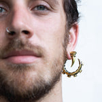 A close-up image of a person with a beard and a nose piercing, wearing the elaborate GIGER Biomechanical Open Ended Hoop Ear Weights in Gold by Kaya Movement. The person's expression is partially obscured, and the background is plain and light. This unique style could easily be found at dystopian festivals or among fans of unisex 0 gauge plug earrings.