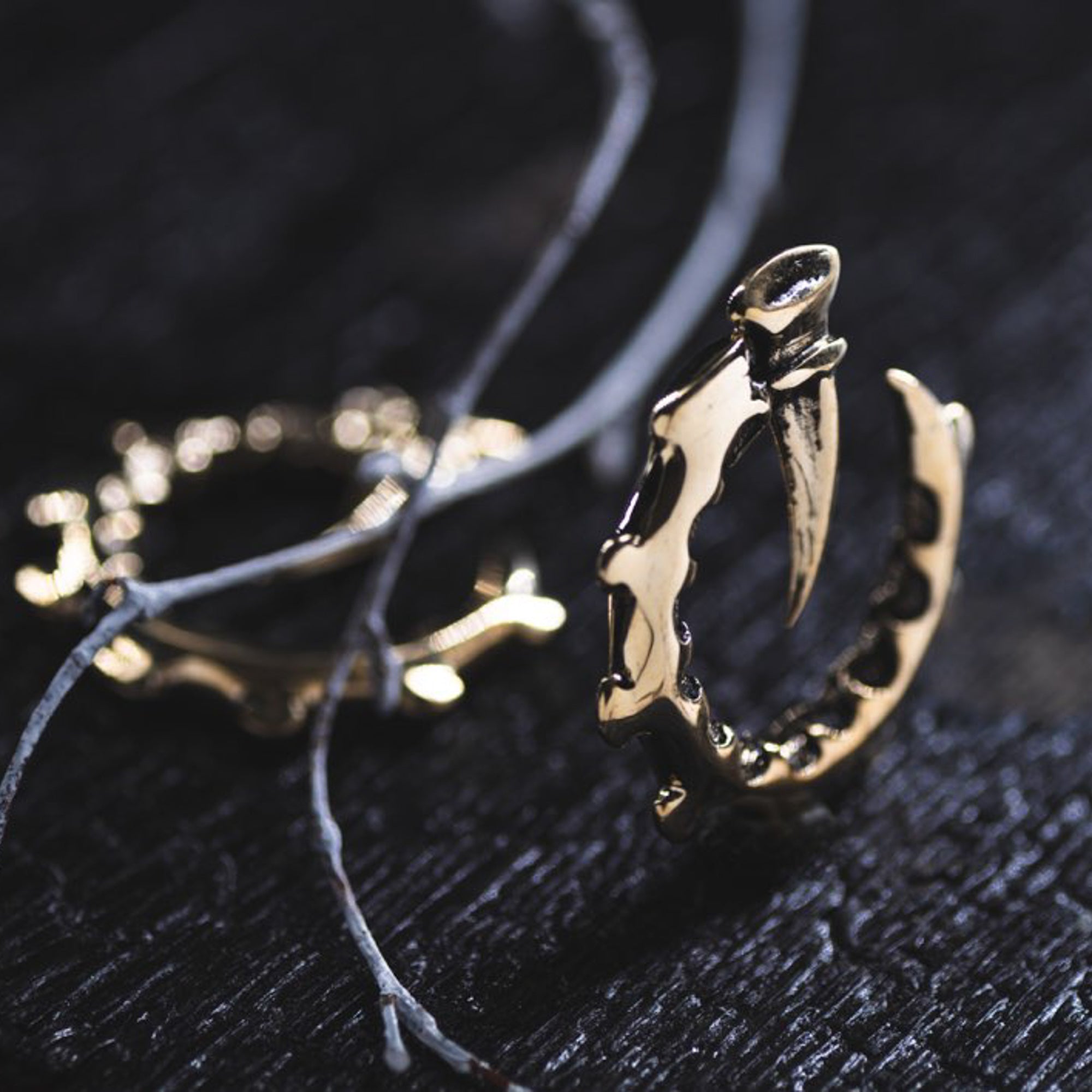 Close-up of the GIGER Biomechanical Open Ended Hoop Ear Weights in Gold by Kaya Movement on a dark, textured surface. Another similar ear weight and some thin, weathered cords are blurred in the background. The ear weights have intricate details, evoking a rugged, edgy aesthetic reminiscent of accessories worn at dystopian festivals.