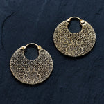 A pair of KONI Tribal Shipibo Endless Hoop Disc Earrings in Gold from Kaya Movement lies on a dark surface. Each round, gold earring features intricate, engraved geometric patterns and a hinged clasp at the top. The symmetrical and detailed design gives them an ornate and elegant appearance, evoking the essence of tribal earrings and the Kaya Movement.