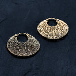 A pair of KONI Tribal Shipibo Endless Hoop Disc Earrings in Gold by Kaya Movement, showcasing a textured design on a dark background. These boho statement earrings feature an 18-gauge circular shape with a small latch closure and display detailed, ornate engravings throughout their surface.