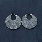 A pair of KONI Tribal Shipibo Endless Hoop Disc Earrings in 925 sterling silver, designed by Kaya Movement, is displayed on a dark textured background. These 18-gauge circular earrings feature elaborate geometric patterns that give them a detailed and artistic appearance reminiscent of tribal body jewelry from the Kaya Movement.