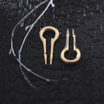 A close-up of two small, golden Kaya Movement CALYPSO Minimalist Keyhole Ear Weights in 2 gauge on a dark, textured surface. The ear weight on the left features a keyhole with two prongs, while the one on the right has two pointed, thinner ends. Thin, white twigs lie beside what resemble post-apocalyptic body jewelry elements.
