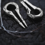 Close-up of two intricately designed EXRO Biomechanical Cyborg Keyhole Ear Weights in Silver by Kaya Movement, resembling silver plier-like tools with a looped handle, set against a dark, textured surface. The ear weights feature ornate, linear patterns on their metallic surface, evoking the elegance of antique post-apocalyptic body jewelry.