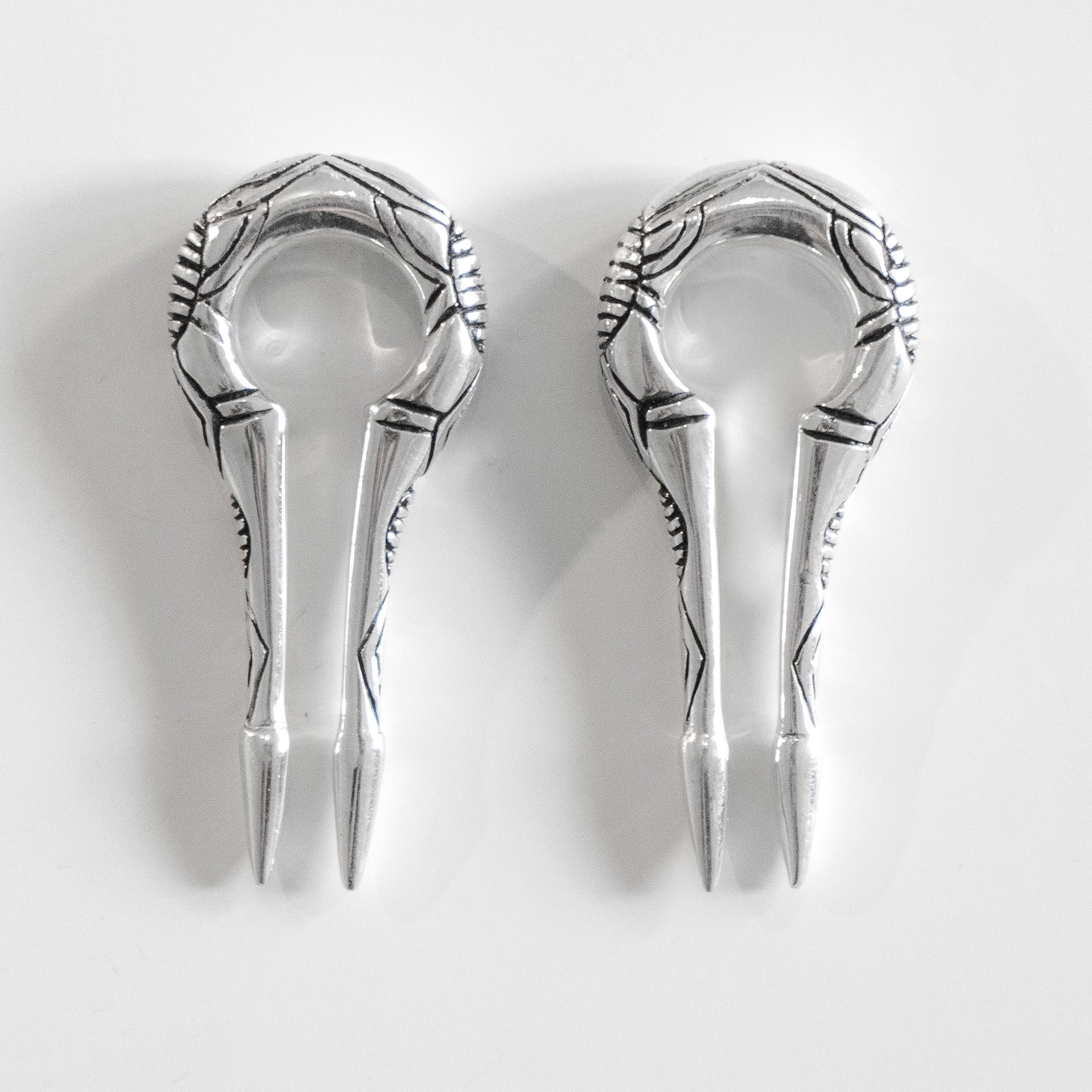 A pair of intricately designed Kaya Movement EXRO Biomechanical Cyborg Keyhole Ear Weights in Silver | 2 gauge, featuring metallic, double-pronged aesthetics with ornamental patterns on a white background. These ear weights have a circular loop at the top and pointed ends, reminiscent of post-apocalyptic body jewelry, showcasing a detailed decorative design.
