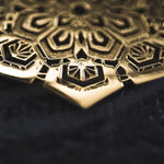A close-up image of the ASA Oversized Asa No Ha Mandala Disc Earrings in Gold by Kaya Movement, showcasing an intricate geometric and floral design reminiscent of ethnic statement jewelry. The lighting highlights the depth and detail of the 16 gauge etched metal, accentuating its metallic sheen against a dark background.