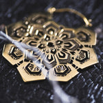 A close-up of a detailed ASA Oversized Asa No Ha Mandala Disc Earring in Gold by Kaya Movement lying on a textured dark surface. The design features intricate geometric patterns and cutouts, reminiscent of ethnic statement earrings, surrounding a central flower-like shape. A blurred white feather is partially visible in the foreground.