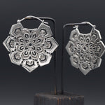 The ASA Oversized Asa No Ha Mandala Disc Earrings in Silver by Kaya Movement are a pair of intricate silver earrings featuring a hexagonal floral and geometric design. These elegant tribal earrings, crafted in a delicate cut-out Asanoha motif, hang from black earring stands against a dark background.