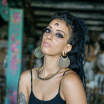 A person with long, dark hair, wearing Kaya Movement's AREVALO Shipibo Oversized Tribal Disc Hoop Earrings in Silver (16 gauge) and a nose ring, gazes at the camera. They have unique facial markings and wear a black top with a gold chain necklace. The background is blurred, giving focus to the subject.