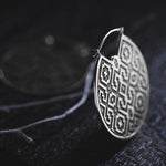 Close-up of the AREVALO Shipibo Oversized Tribal Disc Hoop Earring in Silver by Kaya Movement, featuring a circular design with intricate geometric patterns. Set against a dark, textured background, the detailed engraving on this statement earring is prominent, with its metallic surface subtly reflecting light.