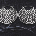 A close-up image of the AREVALO Shipibo Oversized Tribal Disc Hoop Earrings in Silver by Kaya Movement lying on a dark textured surface. The statement earrings, crafted with a 16 gauge silver brass, showcase an intricate geometric pattern featuring symmetrical diamond and cross shapes. Twigs are scattered around them.
