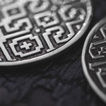 Close-up of AREVALO Shipibo Oversized Tribal Disc Hoop Earrings in Silver by Kaya Movement, reflecting intricate geometric patterns and symbols, placed on a dark, textured surface. The earrings' detailed designs resemble the craftsmanship seen in statement jewelry, showcasing a combination of lines, crosses, and abstract shapes.