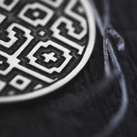 Close-up image of the AREVALO Shipibo Oversized Tribal Disc Hoop Earrings in Silver by Kaya Movement, showcasing their intricate geometric patterns and symbols. The round, metallic earrings lie gracefully on a dark, textured surface. To the right, twisted metal wires or branches blend seamlessly into the composition.