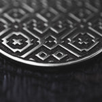 A close-up of the AREVALO Shipibo Oversized Tribal Disc Hoop Earrings in Silver by Kaya Movement, highlighting their intricate geometric pattern with interlocking shapes and stars. The embossed design creates a textured appearance against the dark background, with lighting accentuating the metallic sheen of these 16-gauge statement earrings.