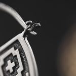 Close-up of the AREVALO Shipibo Oversized Tribal Disc Hoop Earrings in Silver by Kaya Movement, featuring an intricate geometric pattern with a prominent cross design in the center. The background is blurred, drawing attention to the fine details and craftsmanship of these statement earrings.