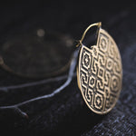 Close-up of a round, intricately patterned earring resting on a dark surface. The AREVALO Shipibo Oversized Disc Hoop Earring in Gold by Kaya Movement showcases sacred geometry Shipibo designs with repeating square and diamond shapes in a gold color. Another similar earring is blurred in the background, enhancing the tribal disc hoop earrings vibe.