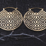 The AREVALO Shipibo Oversized Disc Hoop Earrings in Gold by Kaya Movement feature large, circular brass design etched with intricate Sacred geometry Shipibo patterns. These 14 gauge tribal earrings rest against a textured dark background, showcasing symmetrical shapes and dots that contribute to their bold, culturally-inspired look.