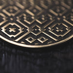 Close-up of the AREVALO Shipibo Oversized Disc Hoop Earrings in Gold by Kaya Movement, featuring intricate geometric and star patterns on their metallic surface reminiscent of Sacred geometry Shipibo designs. The detailed interlocking shapes and fine lines create a textured appearance against a dark, blurred background.