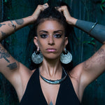 A woman with PANCHA Oversized Tribal Shipibo Disc Earrings in Silver by Kaya Movement and a silver necklace stands with her hands behind her head. She has long black hair, tattoos on her arms, and wears a black halter top. The background is dark with a greenish hue, possibly a mossy or natural setting.