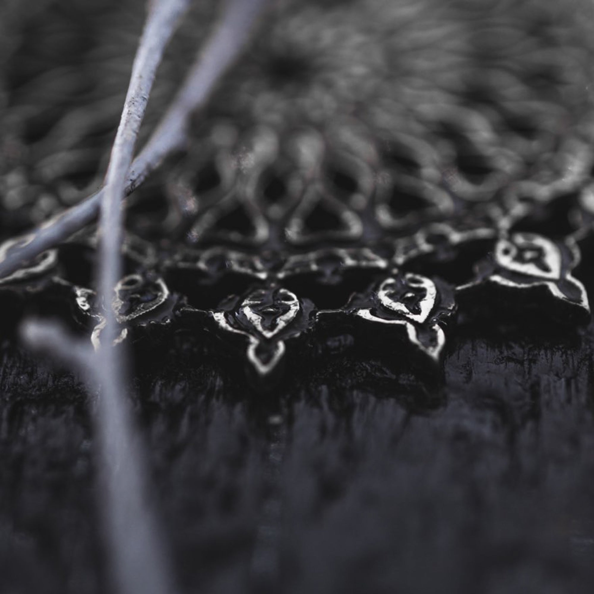 Close-up of the intricate black and silver metallic pattern on the MOZAIKA Oversized Gothic Disc Earrings in Silver by Kaya Movement, showcasing detailed filigree work reminiscent of classic Gothic designs. Thin silver threads are draped across the foreground, slightly out of focus. The dark background enhances the contrast and details, making it a striking visual.