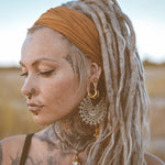 A person with long dreadlocks tied back with a brown headband gazes downward. They have multiple facial piercings, including a septum piercing, and wear large, intricate Mozika Oversized Gothic Disc Earrings in Gold Brass by Kaya Movement. Their face and neck are adorned with various tattoos. The background is blurred and natural.