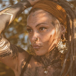A person with braided hair adorned with a headband gazes into the distance. They have multiple facial piercings, large intricate Kaya Movement Mozika Oversized Gothic Disc Earrings in Gold Brass (16 gauge), and full arm tattoos. Dappled sunlight creates a patterned shadow across their face, set against a backdrop of outdoor foliage.