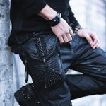 A person clad in black pants adorned with intricate leather and metal details leans casually against a wall. Their ensemble features a black wrist accessory, studded black boots, and the Kaya Movement's APOPHIS Post-Apocalyptic Leather Shoulder & Belt Bag with Black Studs slung across their chest. The detailed design of the pants and the person's relaxed stance are highlighted.
