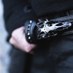 A close-up image of a person wearing an all-black outfit highlights the APOPHIS Post-Apocalyptic Leather Shoulder & Belt Bag by Kaya Movement, adorned with black studs, making it an ideal accessory for your next wasteland festival. The person's hand is partially visible, resting on or near the belt bag. The background is blurred to emphasize the unique details of the bag.