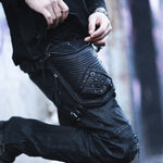 A person wearing black cargo pants with distressed details and a wasteland festival accessory, specifically the APOPHIS Post-Apocalyptic Leather Shoulder & Belt Bag by Kaya Movement, complete with metal embellishments, attached to the thigh. They are adjusting the strap of this utility piece, with only their midsection and hands visible.
