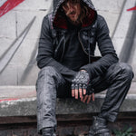 A person wearing the VENOM Men's Leather Hooded Vest Jacket by Kaya Movement, paired with black pants and fingerless studded gloves, sits on a concrete ledge. Their long hair partially covers their face, set against a backdrop of a rough, graffiti-marked wall. The ensemble screams rebellion with hints of punk rock biker jacket vibes.